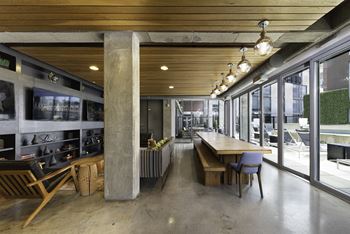 A long wooden table with chairs is in the middle of a room with large windows